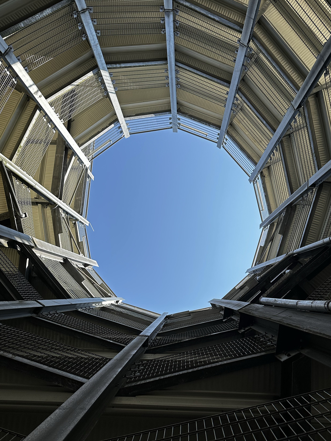 View from the bottom of a circular metal structure with mesh panels and support beams, looking up at a clear blue sky through an open top. The perspective creates a sense of depth and symmetry.