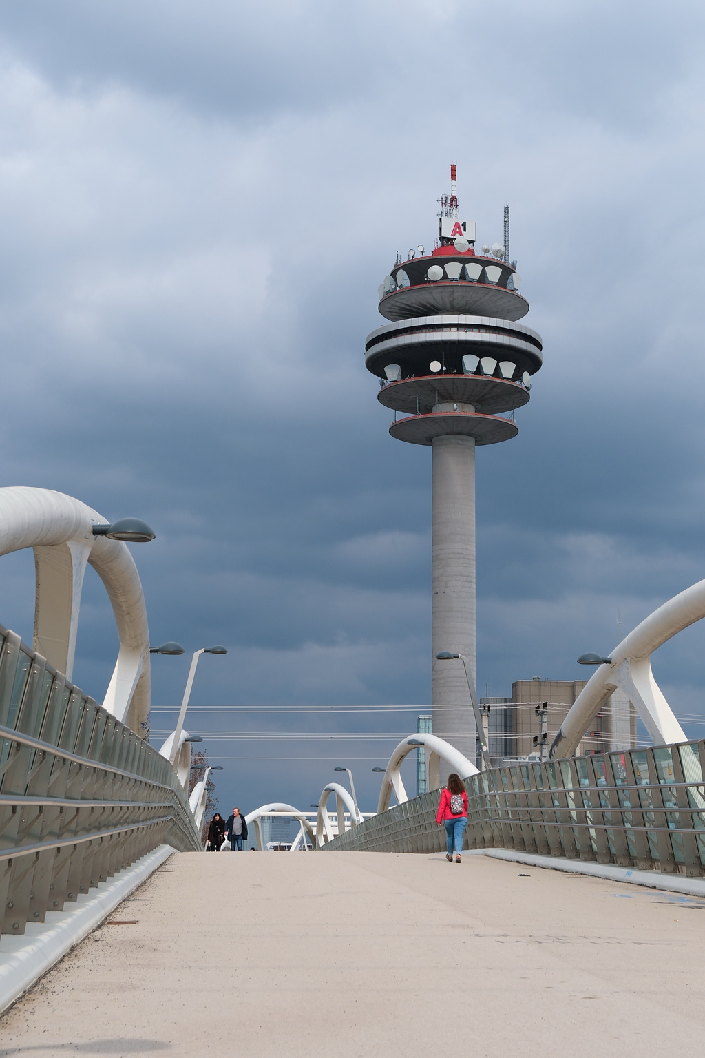 A person wearing a red jacket walks across a modern, arched pedestrian bridge. In the background, a tall telecommunications tower with multiple circular dishes is visible against a cloudy sky. Several street lamps line the bridge, and a glass railing runs along the sides.
