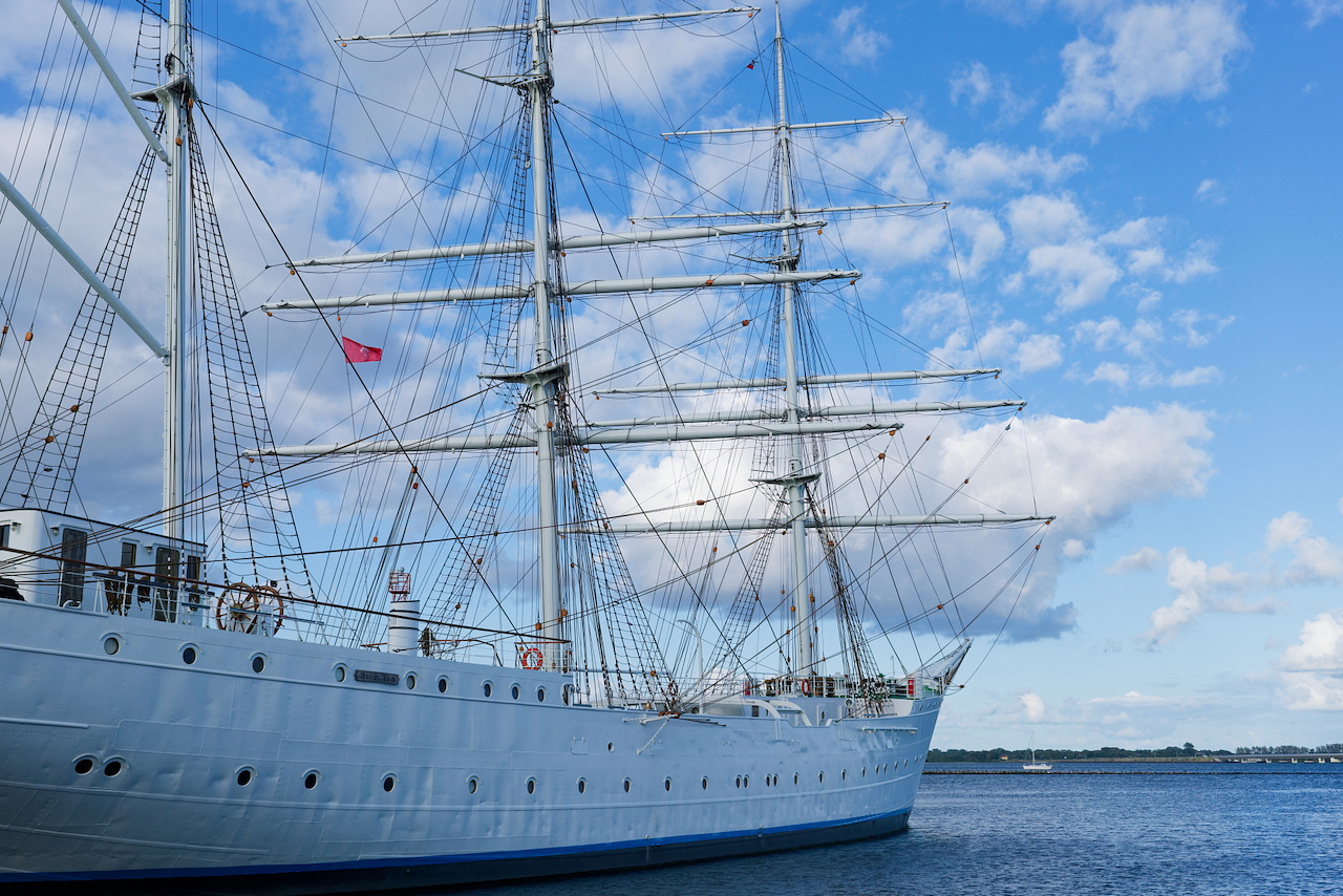 A large white sailing ship, the Gorch Fock, with multiple masts and rigging is docked in calm water. The sky is blue with scattered clouds, and a small red flag is visible on one of the masts. The scene conveys a peaceful maritime atmosphere.