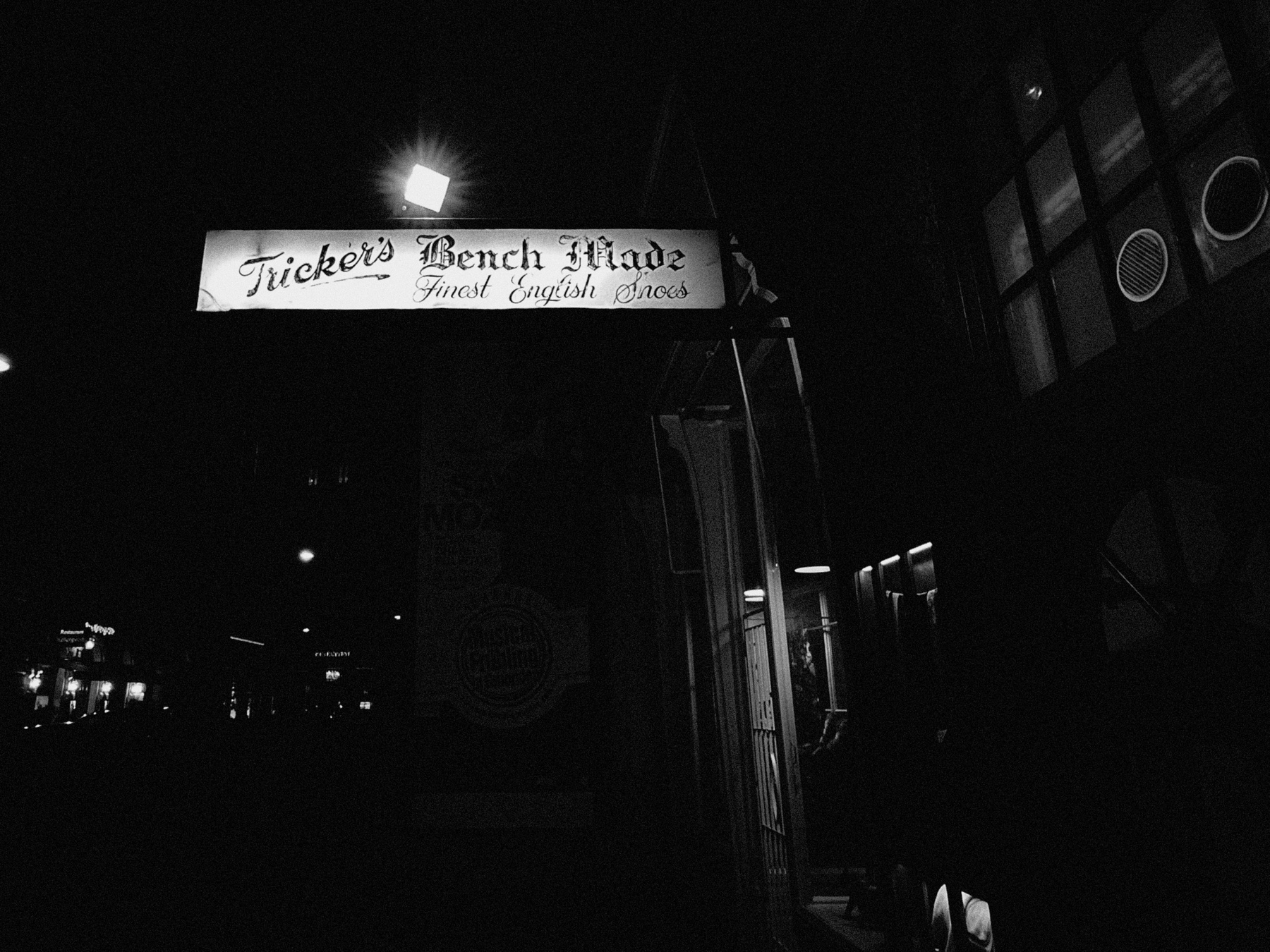 A dimly lit black and white image of a city street at night. The primary focus is on an illuminated sign reading "Tricker's Bench Made, Finest English Shoes." The surrounding area is dark, with faint outlines of buildings and streetlights visible in the background.
