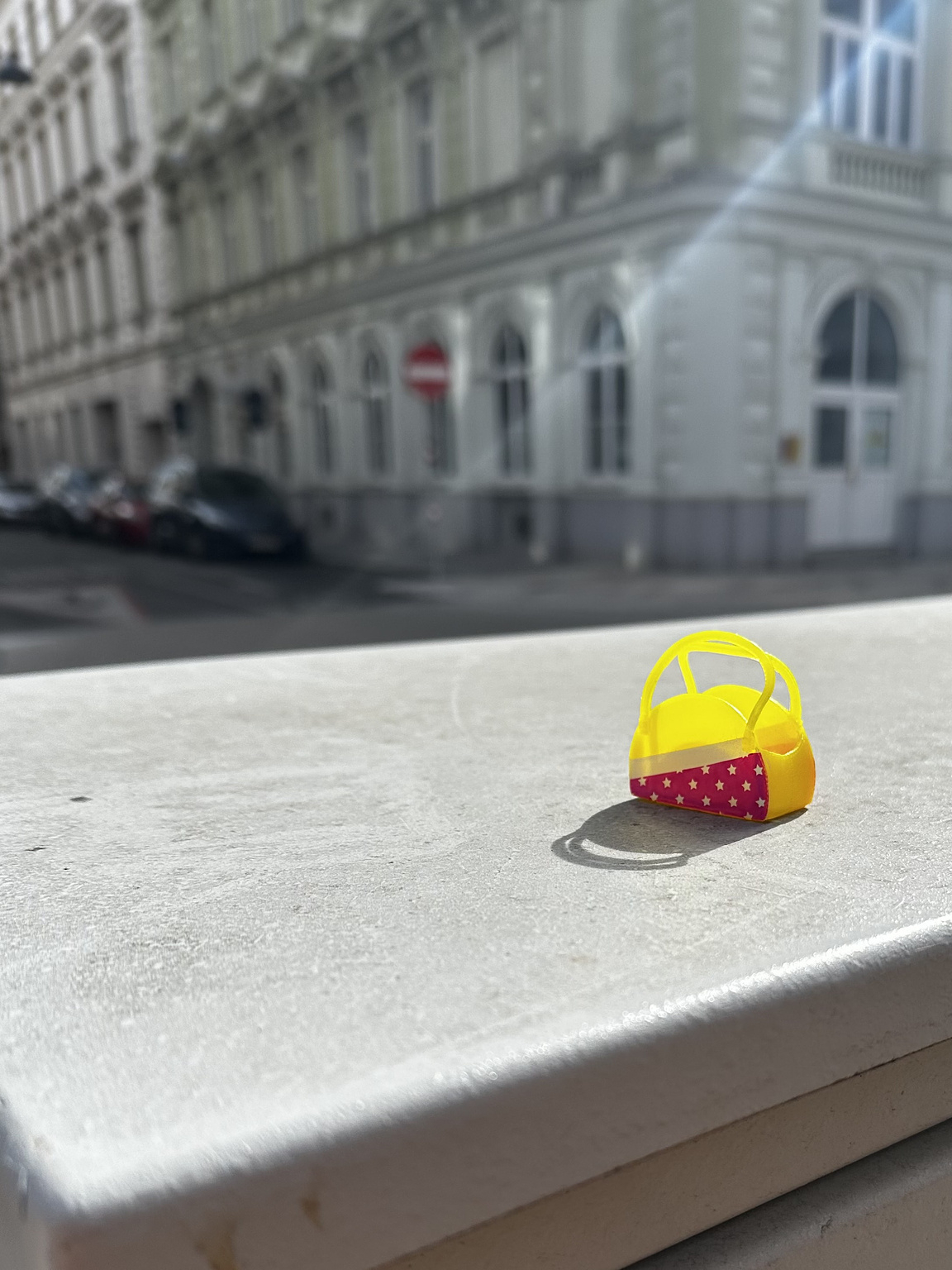 A small, colorful toy basket with a yellow top and pink sides featuring star patterns sits on a ledge. The background shows a blurred view of a street with an ornate building, featuring arched windows. Sunlight creates a bright highlight on the toy and casts its shadow onto the ledge.