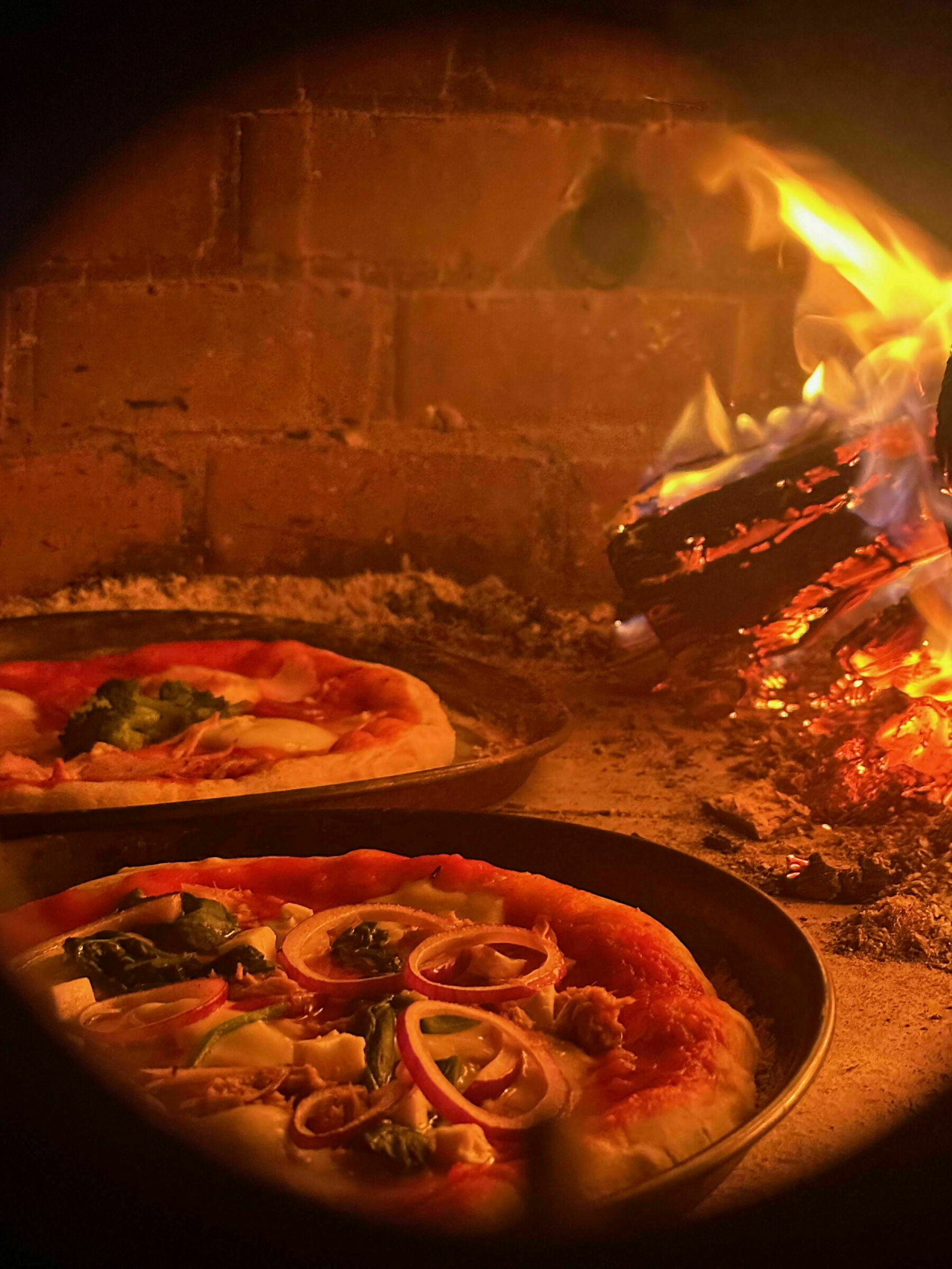 Two pizzas in a pizza oven with a wood fire burning inside.