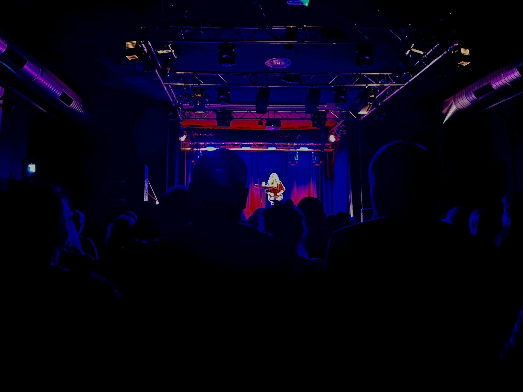 A dark hall in an event center with people sitting on chairs in front of a stage on which a woman sits on a bar stool at a small table to give a reading.