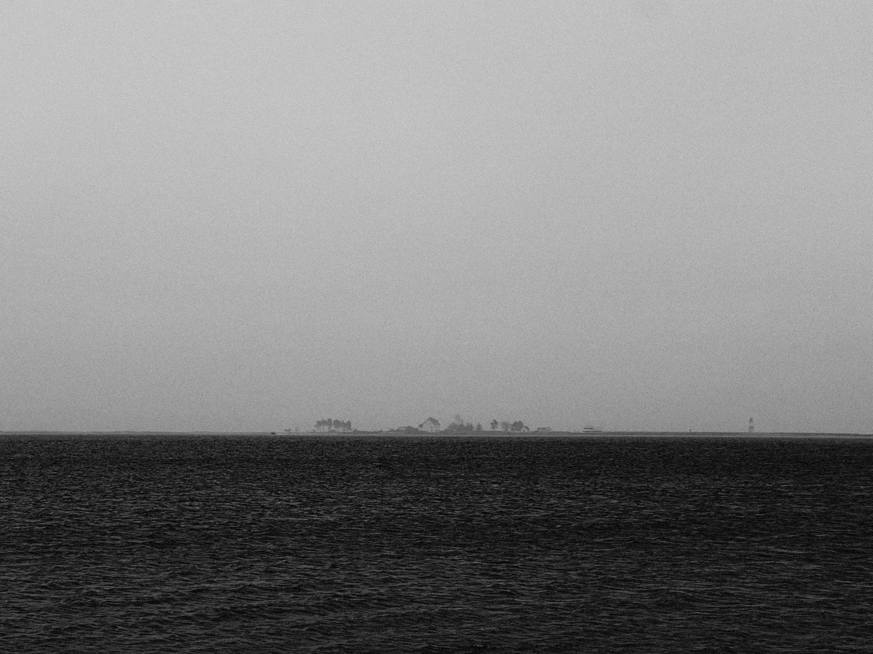 A black and white photograph of a distant shoreline, featuring a small, faint island with trees barely visible on the horizon. The foreground is dominated by a vast, dark sea with gentle ripples. The sky is overcast and merges smoothly with the water, creating a minimalist and atmospheric scene.