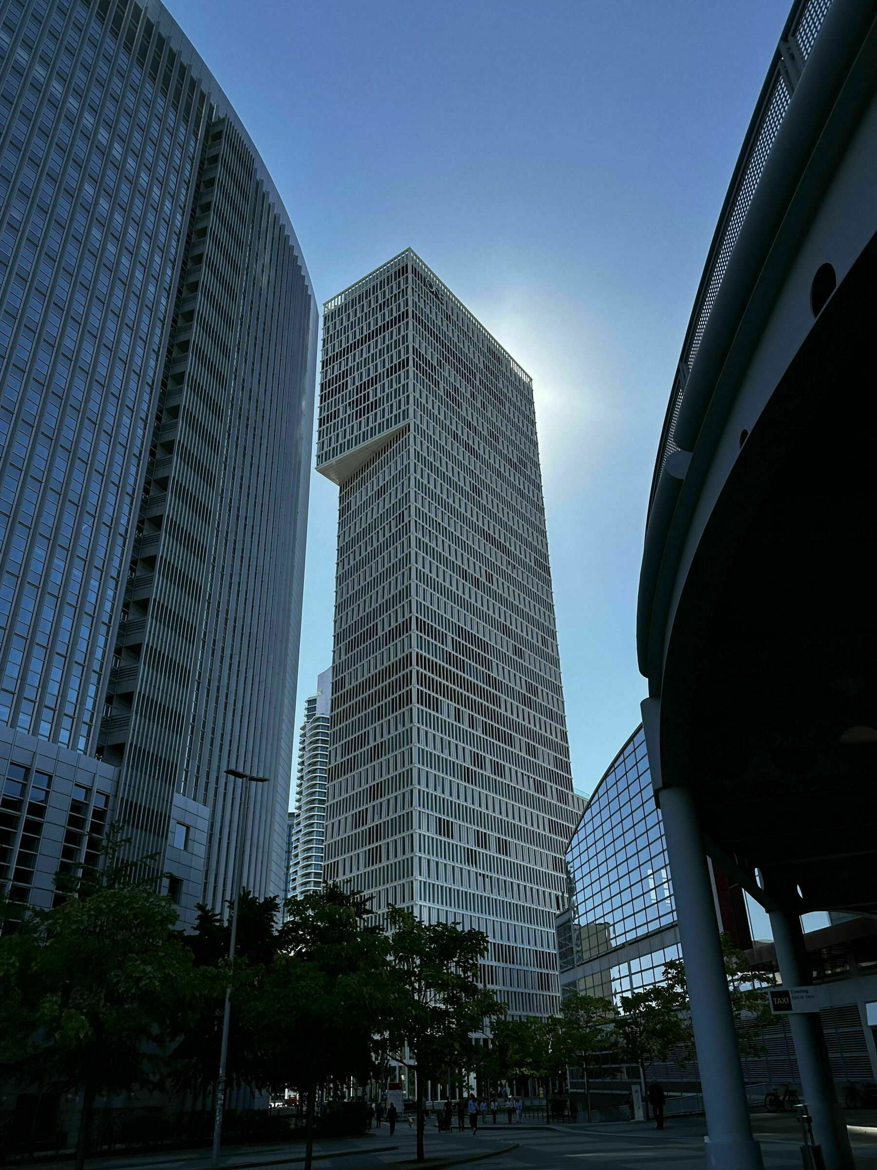 Tall modern buildings against a clear blue sky. The central skyscraper has a distinctive overhanging section near the top, casting a bright silhouette with the sun positioned directly behind it. Surrounding the buildings are several trees, and there’s a curved, glass-covered structure on the right. Shadows create contrasting patterns on the ground.