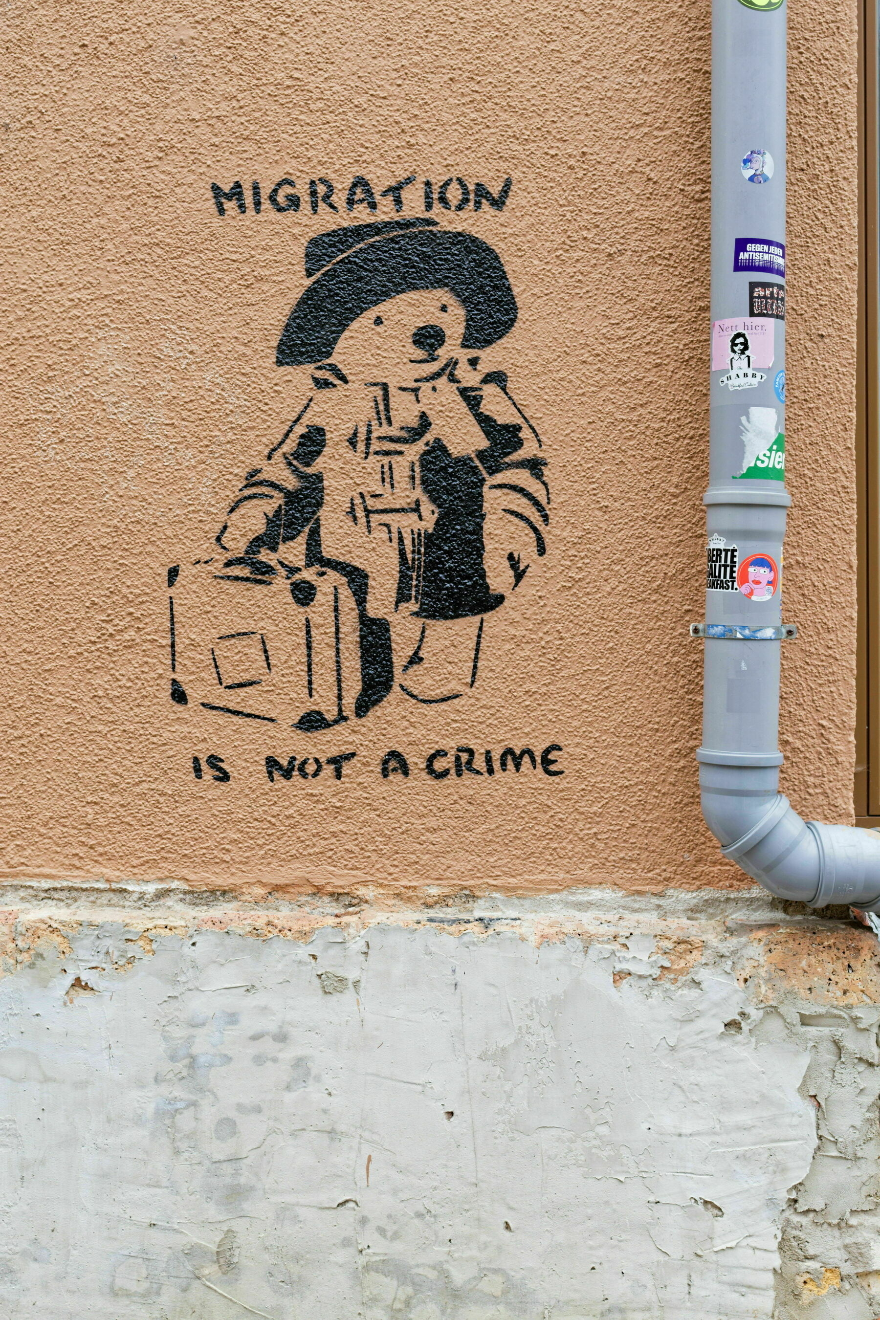 Street art on a textured wall shows a figure of a bear wearing a coat and hat, holding a suitcase. The text above and below reads