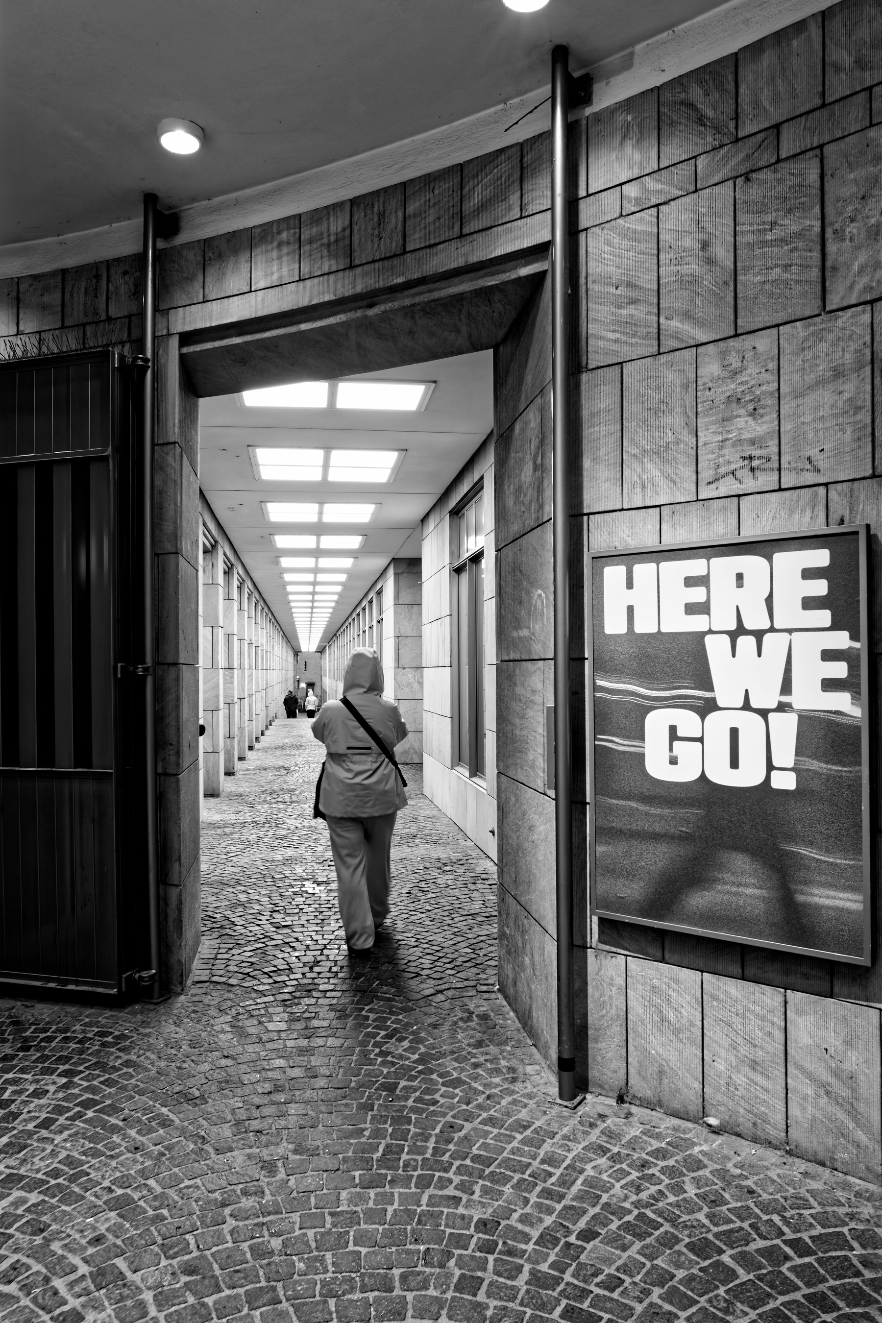 A black and white photograph of a narrow cobblestone passageway lined with tiled walls and overhead lighting. A person wearing a hooded jacket is walking away from the camera. To the right, there's a large sign with the words