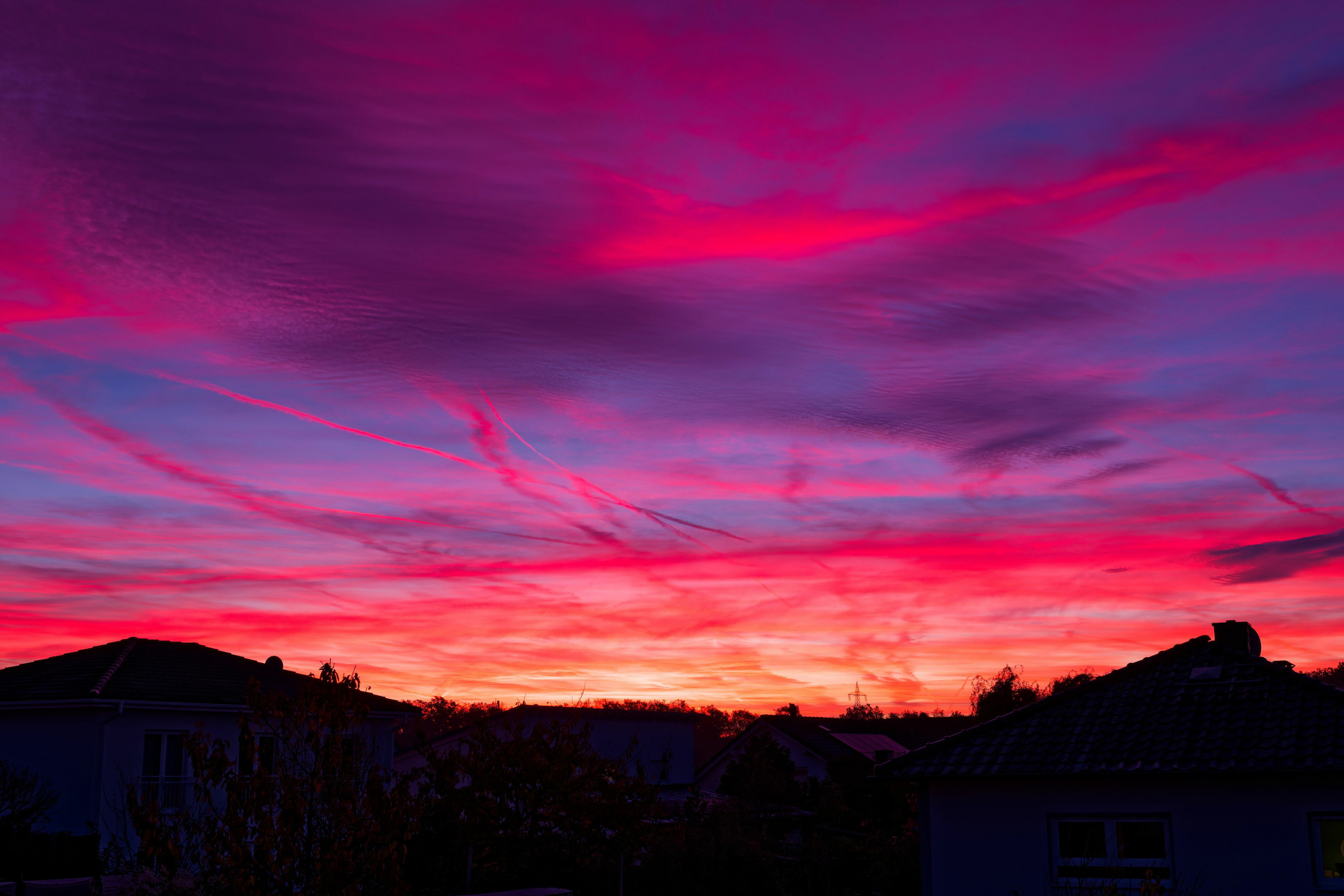 A dramatic sunset with streaks of pink and red clouds dominating the sky. Silhouetted rooftops are visible along the horizon.