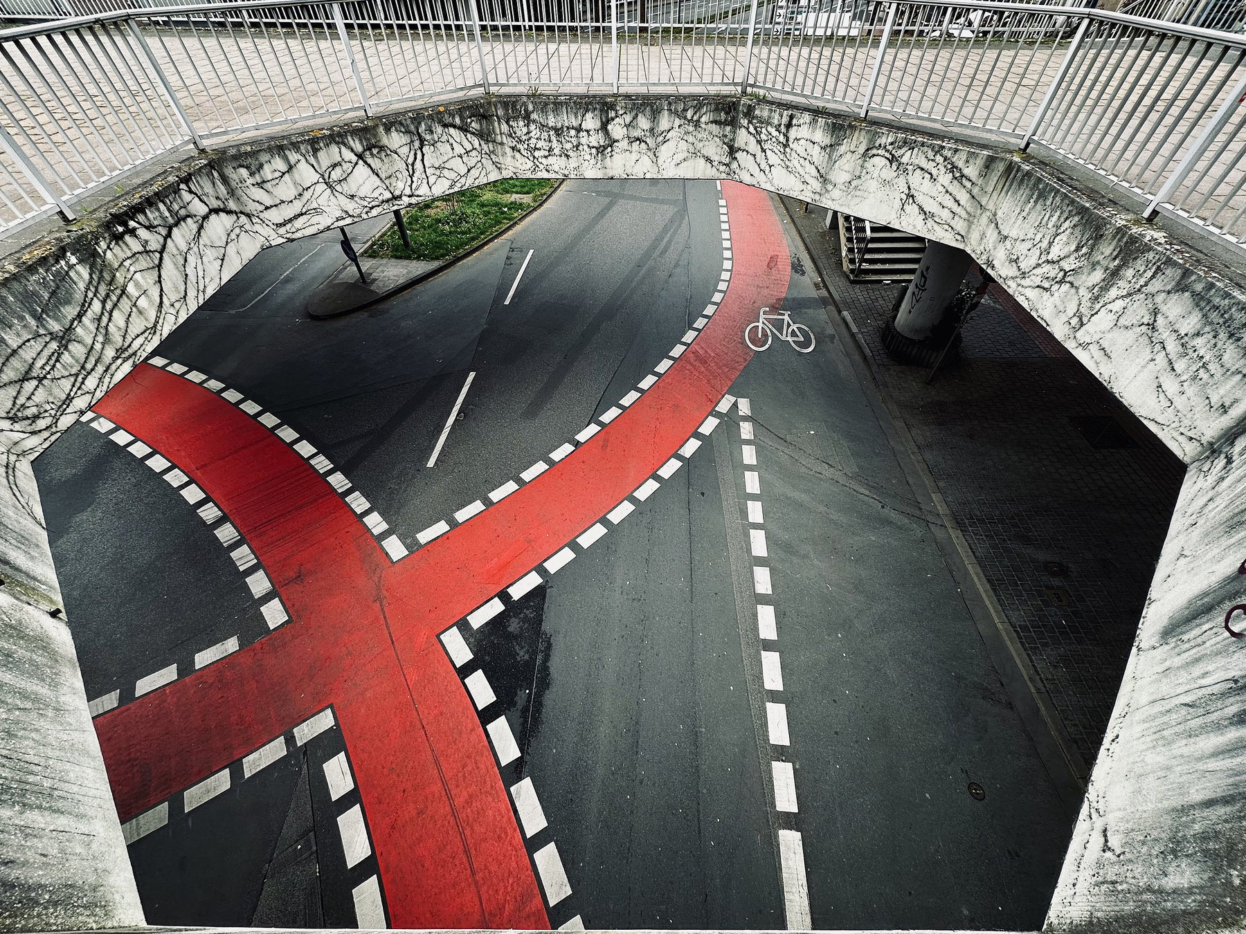 View from a pedestrian bridge showing a road intersection. A red, X-shaped bike lane is marked distinctly, with dashed white lines bordering it. A white bicycle symbol is painted on the bike lane indicating its purpose. Surrounding roads have marked lanes, and green vegetation is visible on a small traffic island. The bridge has a textured, aged appearance with metal railings.