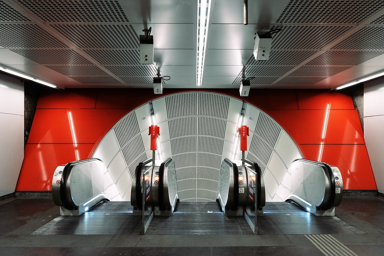 A modern indoor escalator setup in a metro or subway station. The design includes two parallel escalators with sleek metallic sides, bright red stop signs, and a contrasting backdrop of red and gray panels. The ceiling has a grid-like pattern, and the ambiance is well-lit with a futuristic aesthetic.