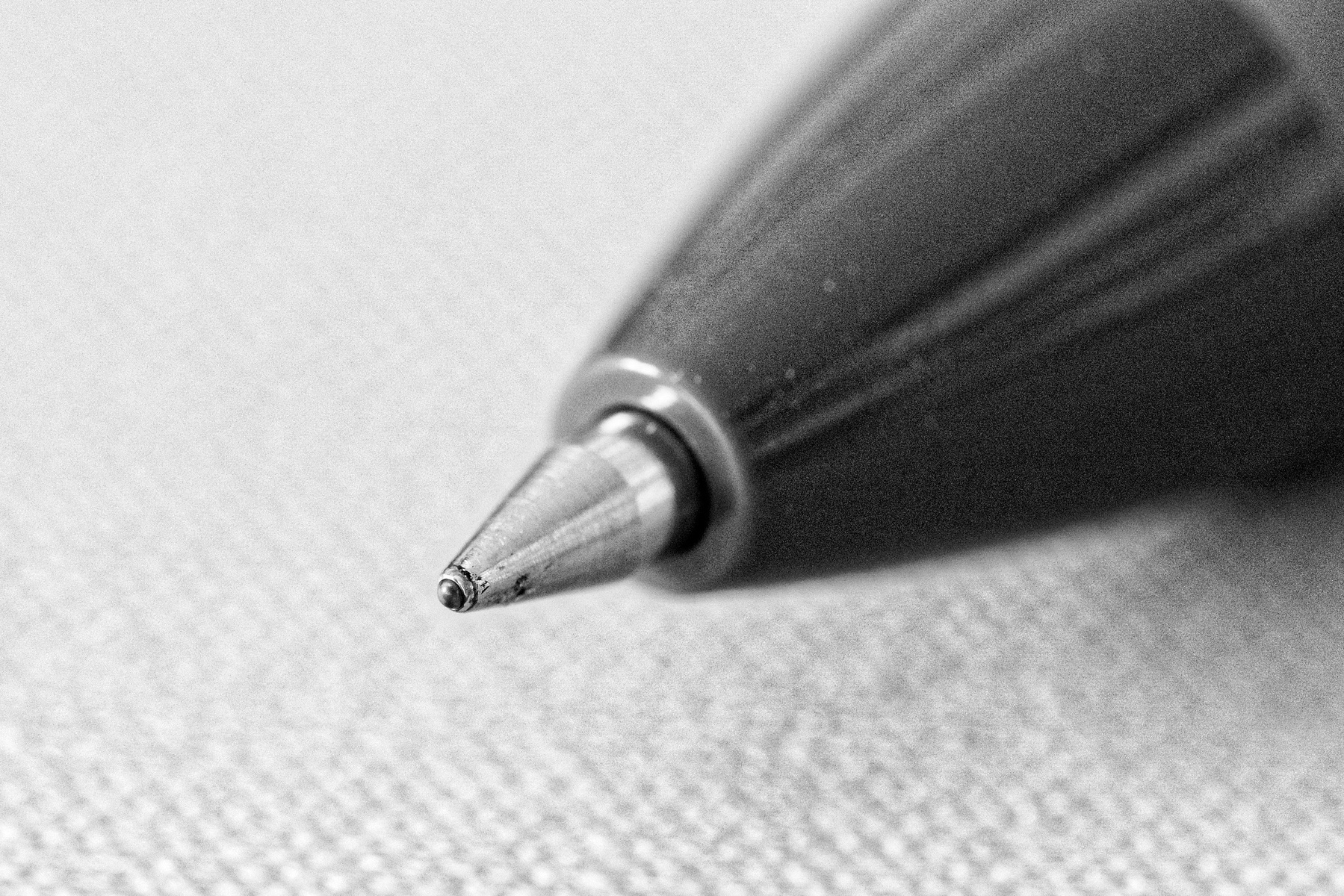 Close-up of a ballpoint pen tip on a textured surface. The pen is in focus, and the image is in black and white, highlighting the metallic tip and plastic casing.