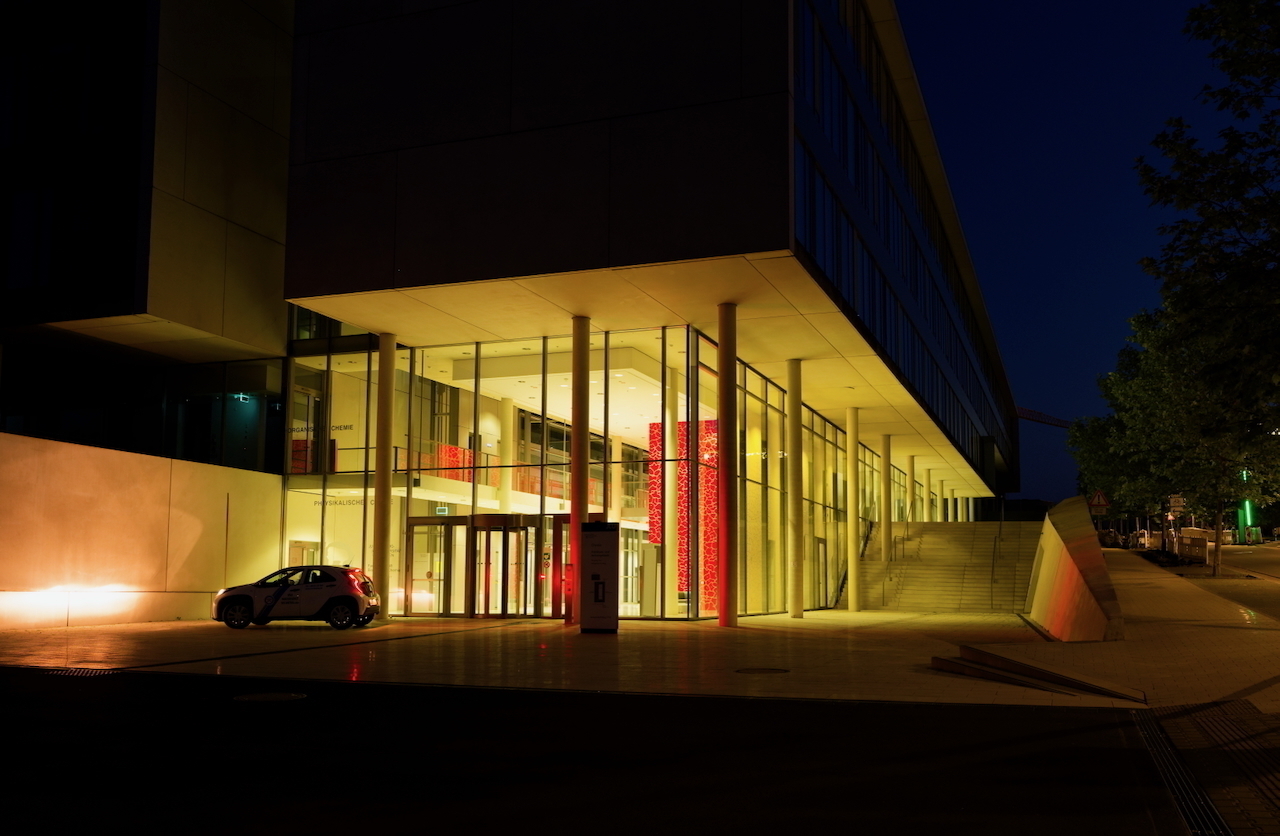 A modern building with large glass windows is illuminated at night, with a car parked near the entrance.