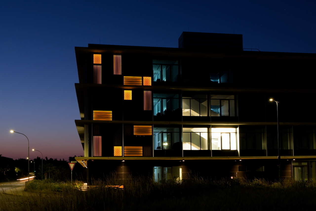 A modern, multi-story building is illuminated at night with geometric lighting design on its facade against a twilight sky.