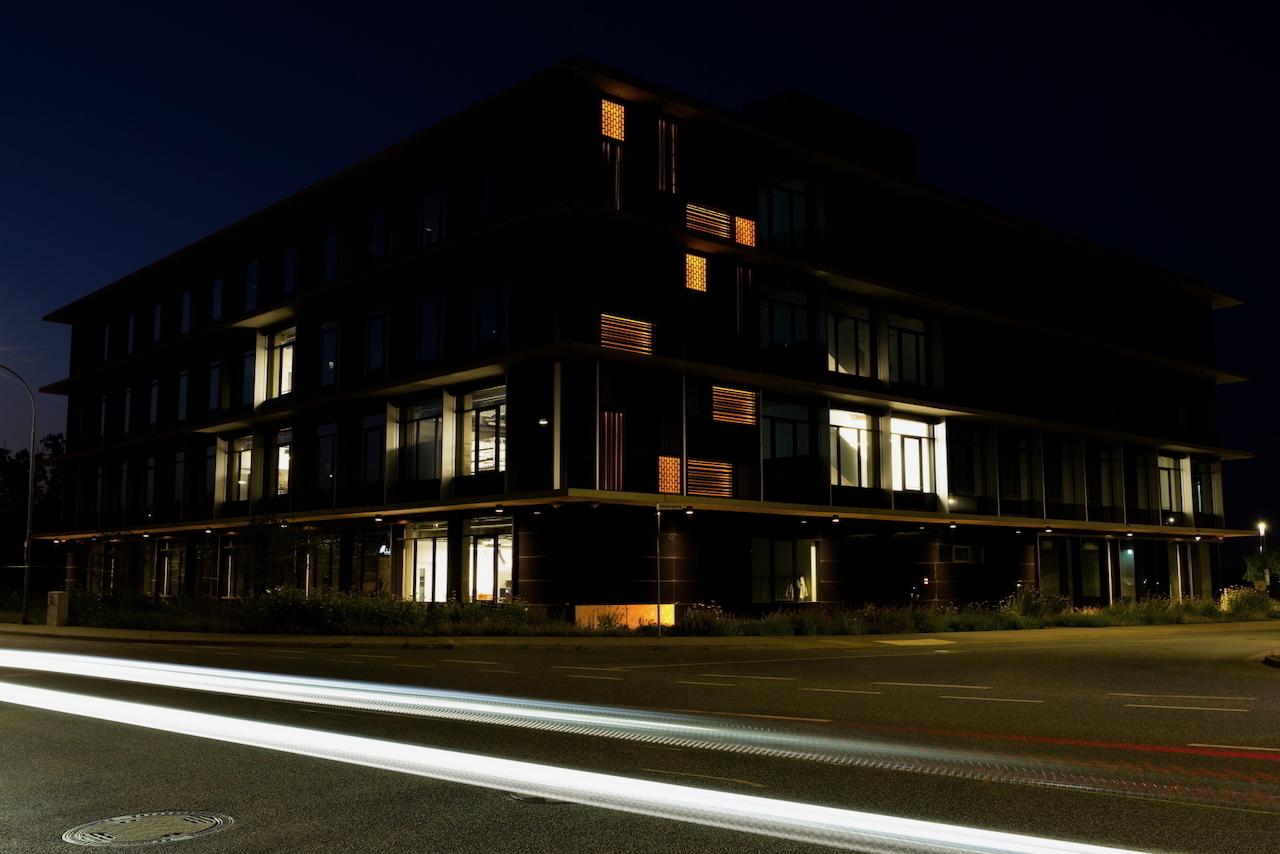 A modern, multi-story building is illuminated in the night, with streaks of light from passing vehicles visible in the foreground.
