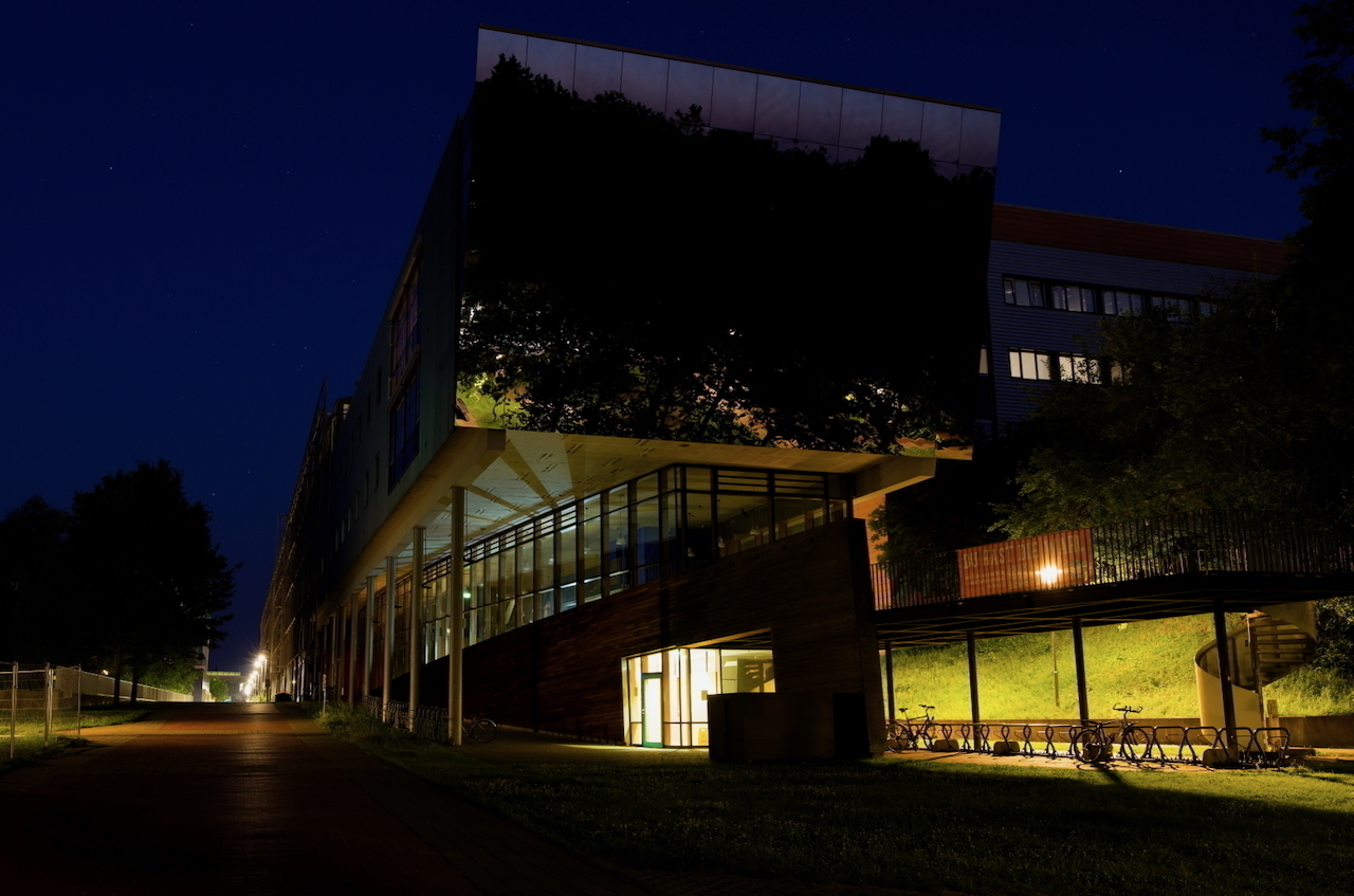 A modern building with large windows is illuminated at night, surrounded by trees and a pathway.