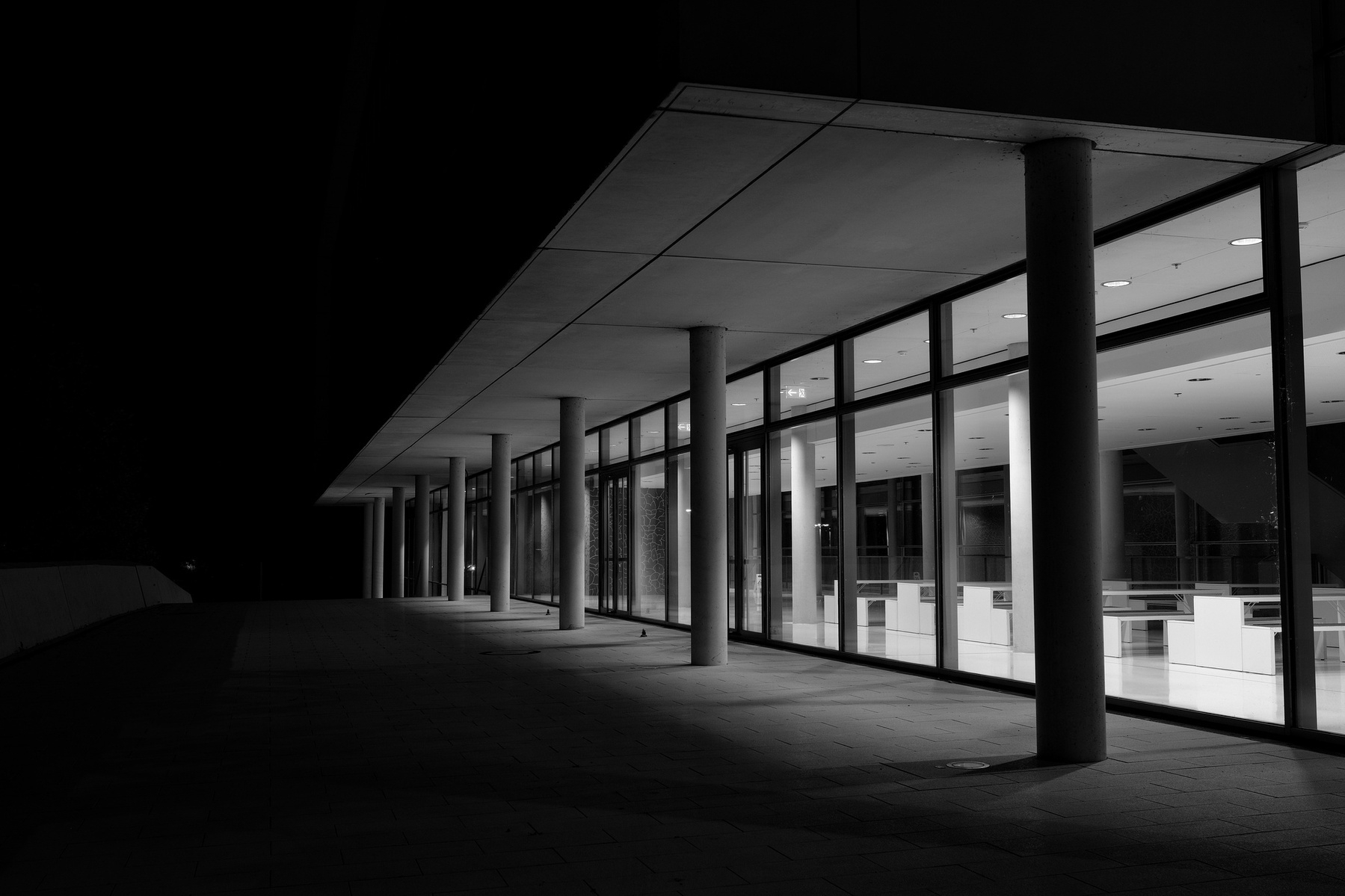 Black and white image of a modern building exterior at night. The structure features large glass windows and a flat roof supported by evenly spaced columns. The interior is well-lit, contrasting with the darkness outside.