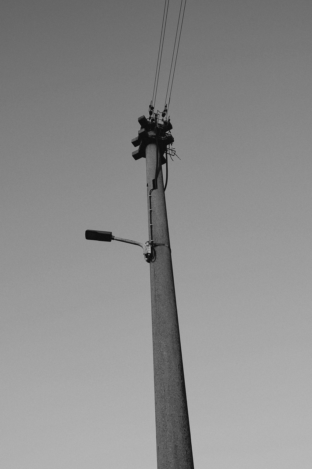 Black and white image of a streetlight pole with a single lamp attached near the top. Power lines extend from the top of the pole, set against a clear sky.