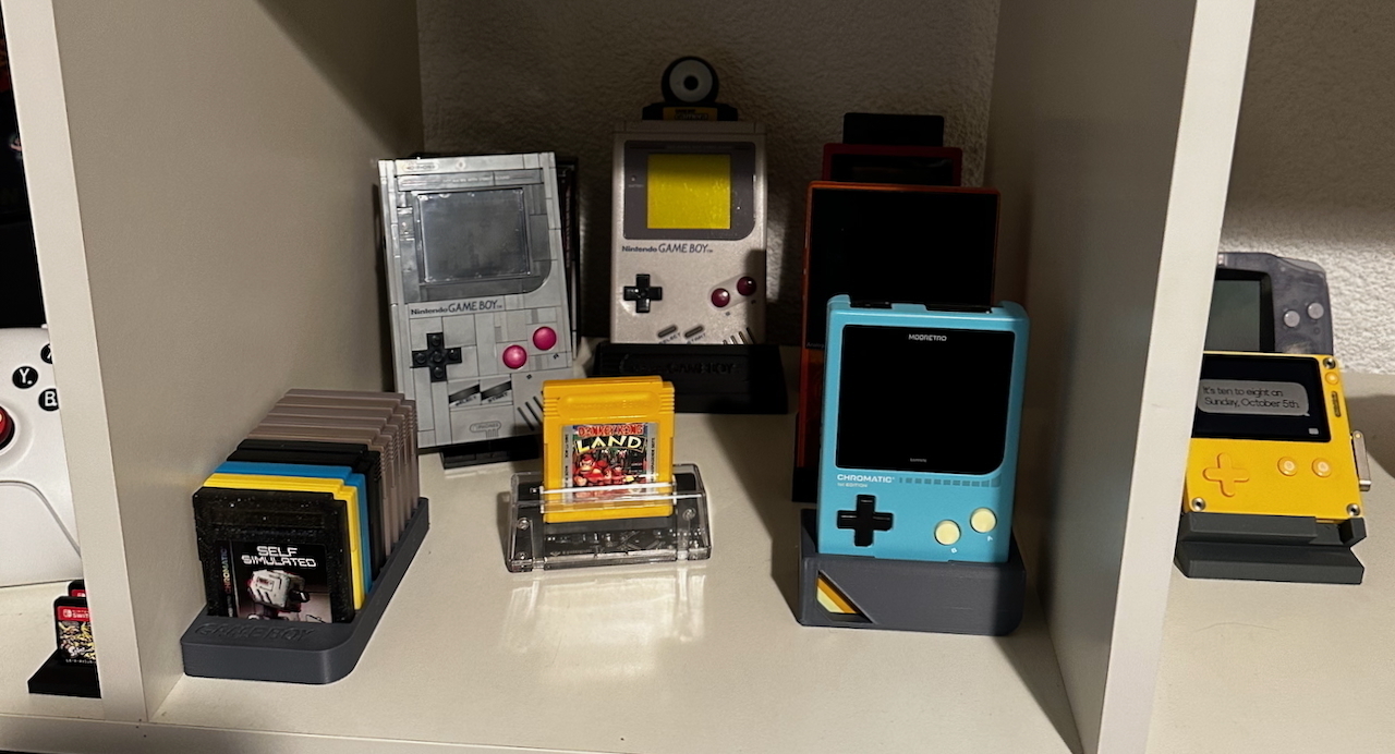 A shelf displaying various handheld gaming devices and cartridges. There are several Game Boys (DMG, Color), a Modretro Chromatic and an Analogue Pocket. Cartridges are neatly arranged, with one prominently featuring "Donkeykong Land" game in a yellow case inside an Epilogue GP Operator. A yellow handheld console with a crank is also visible, a Playdate device.