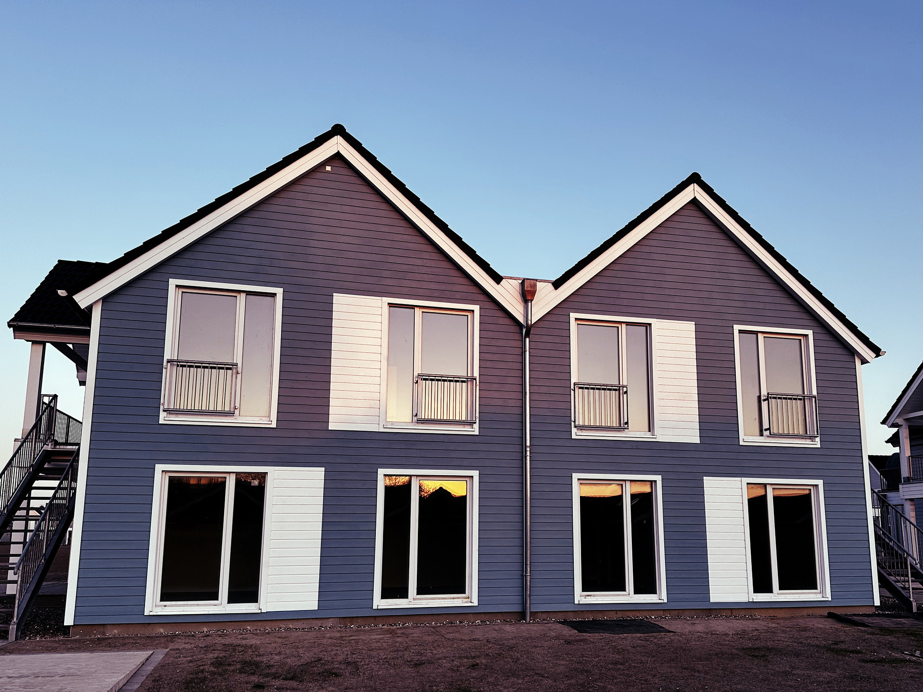 A modern, two-story house with blue siding and white trim. The roof is peaked, and the facade has several large windows. There are two visible exterior staircases on either side of the building. The sky is clear and blue, suggesting a calm weather setting.