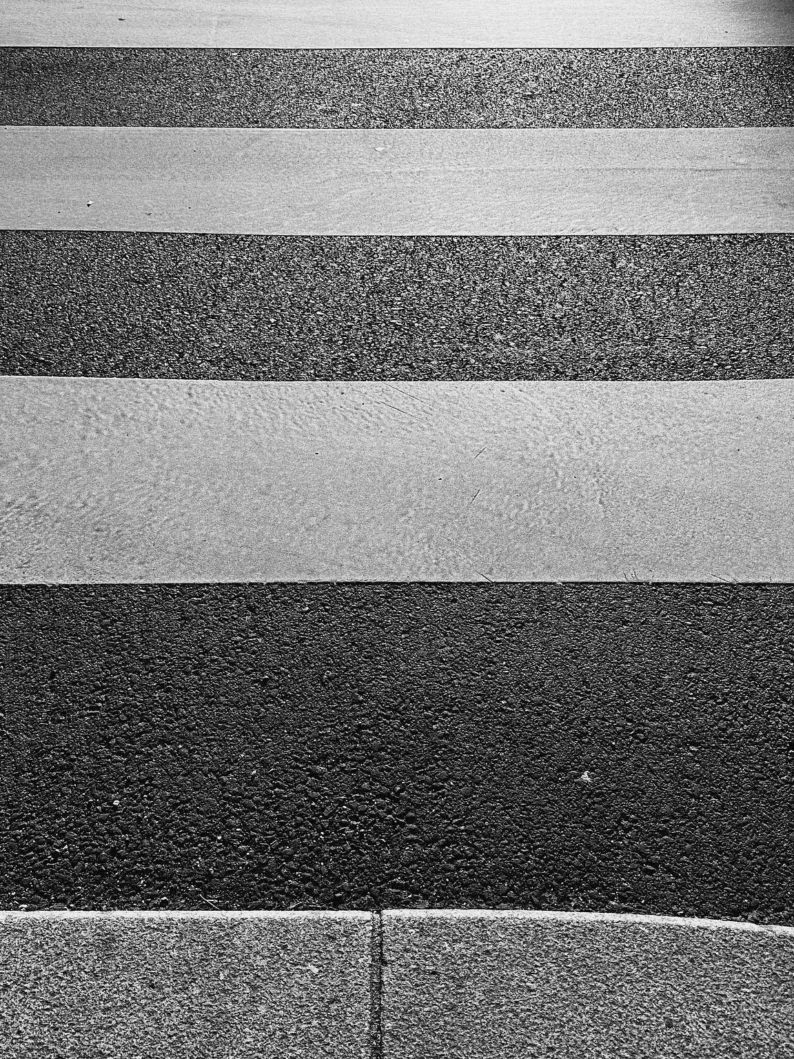 Black and white image of a pedestrian crosswalk, featuring alternating light and dark textured stripes. The surface appears rough, typical of outdoor pavement, with a curved edge of a sidewalk visible at the bottom.